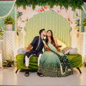 Candid photo of an Indian bride in a dark green lehenga and the groom in a navy kurta sharing a kiss on the cheek, seated on a green couch against a mint-green and floral-themed wedding stage.